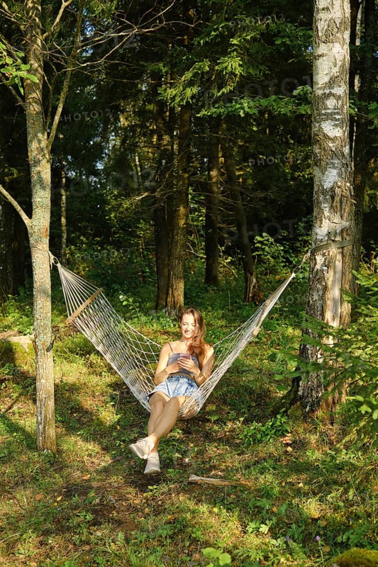 A relaxed woman reads message on smartphone while lounging in a hammock amidst lush green woods