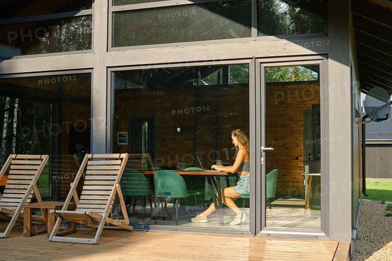 A woman is inside a modern country house with large windows and watches a TV series on a laptop while sitting at a large dining table