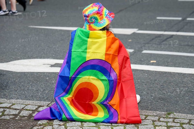 A participant with a rainbow flag and colourful hat sits on the side of the road at the 47th Christopher Street Day under the motto Never be silent again in Berlin on 26.07.2025