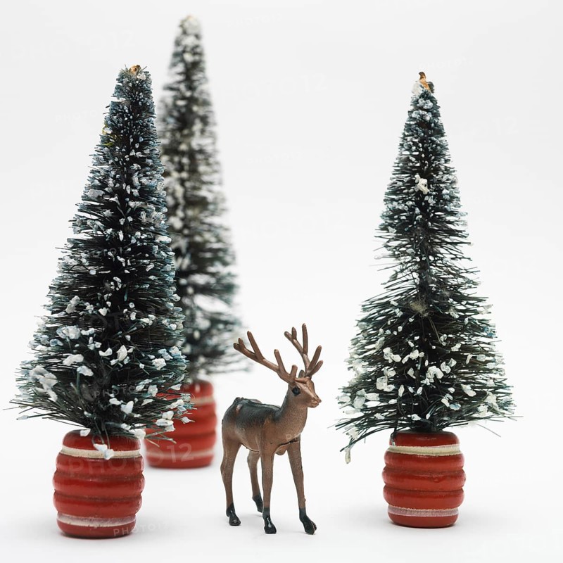 A detailed deer figurine stands in front of two snowy miniature trees, creating a festive atmosphere. This charming arrangement enhances holiday decorations and celebrations