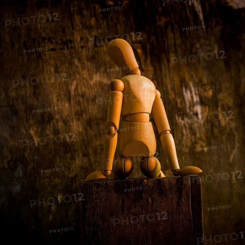 A wooden mannequin is positioned on a rough wooden surface, looking contemplative against a textured, dimly lit background. The warm tones enhance the somber mood of the scene