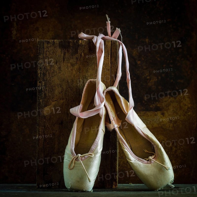 Delicate ballet shoes are placed on a wooden block, showcasing their soft texture and ribbons. The warm brown background enhances their classic beauty and significance in dance