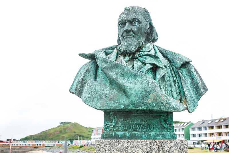 Close-up of the sculpture, monument, bust, bronze bust of August Heinrich Hoffmann von Fallersleben, author of the later German national anthem, The Song of the Germans, on a stone pedestal with a view of the sea in the background the lobster shacks, colourful, brightly coloured buildings, writer, poet, unity and justice and freedom, spa promenade Helgoland, Schleswig-Holstein, Germany