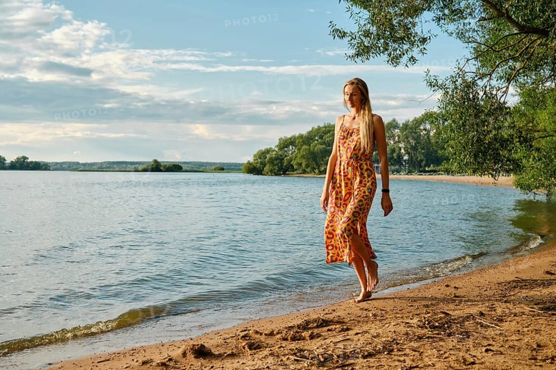 A woman in a colourful sundress strolls barefoot along the sandy shore of a tranquil lake. Lush green trees frame the water under a bright blue sky with fluffy clouds
