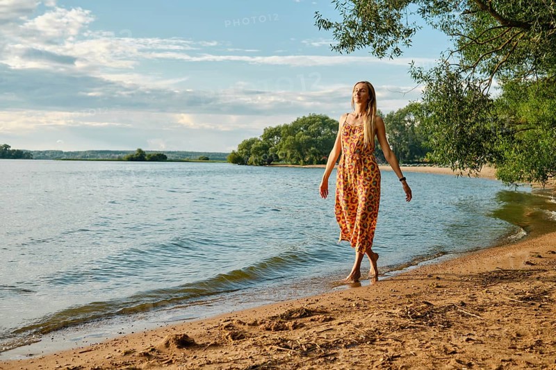 A woman in a vibrant sundress strolls barefoot along the sandy beach beside a calm lake. Sunshine illuminates the landscape, creating a peaceful atmosphere surrounded by lush greenery
