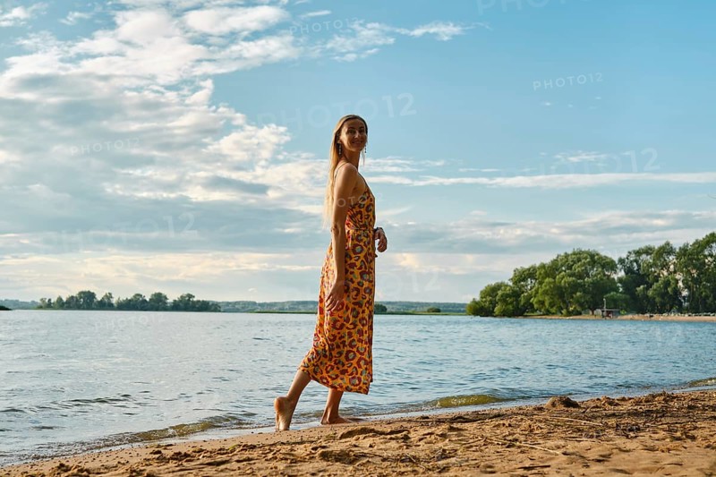 A woman is strolling barefoot along a sandy riverbank, wearing a colourful floral sundress. The blue sky and soft clouds create a serene atmosphere as the sun sets over the water