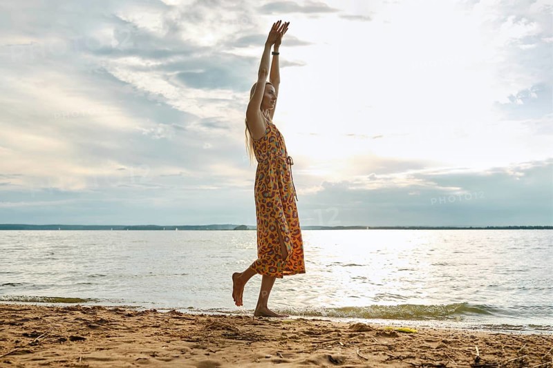 A woman in a colourful sundress walks along a serene lake, raising her arms in joy. The cloudy sky casts a soft light as waves gently lap at the sandy shore, creating a tranquil atmosphere