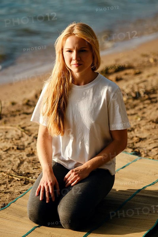 A young woman sits cross-legged on a mat at the beach, gazing thoughtfully towards the water. The calm waves reflect the soft light of the rising sun, creating a tranquil atmosphere