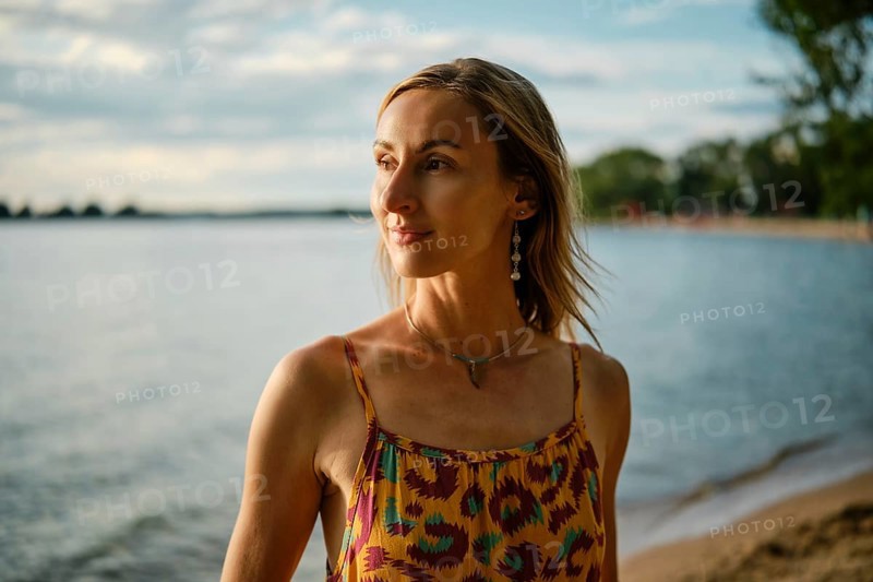 A woman stands near the water's edge, basking in the warm glow of sunset. She wears a colorful dress and reflects peacefully while enjoying the tranquil lakeside atmosphere
