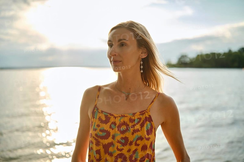 A woman with long hair poses by a calm body of water during sunset, wearing a vibrant patterned dress. Sunlight glimmers on the surface, creating a tranquil and captivating ambiance