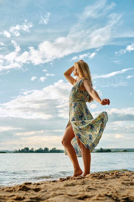 A woman stands gracefully on the sandy shore, her floral dress flowing in the wind. She gazes thoughtfully at the lake under a bright sky filled with clouds, creating a serene atmosphere