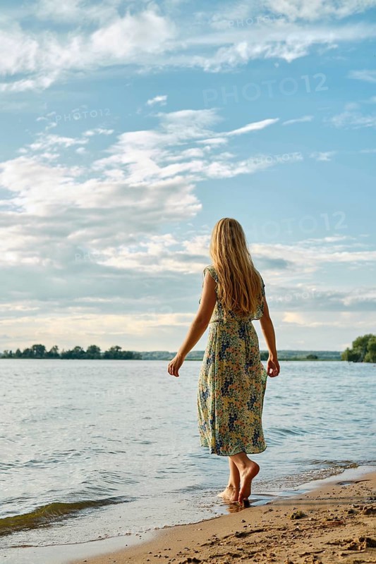 Rear view of barefoot woman standing on a sandy beach by a tranquil lake. The clear blue sky features scattered clouds, creating a serene atmosphere. Time appears to be around late afternoon