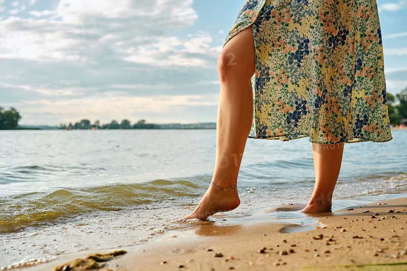 Unrecognizable woman stands at the edge of a lake, her bare foot touching the gentle waves at sunset. She wears a flowery dress, and the sun casts a warm glow on the serene landscape