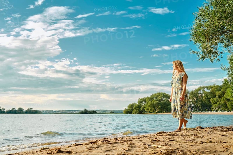 A woman in a floral dress strolls barefoot along the sandy shore of a peaceful lake, surrounded by lush green trees. The clear blue sky and scattered clouds create a tranquil atmosphere