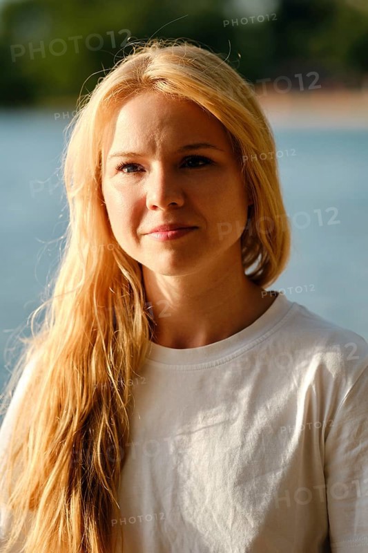 A woman with long flowing hair stands by a serene body of water. Her relaxed expression showcases joy in the sunlight as she enjoys a peaceful afternoon outdoors surrounded by nature