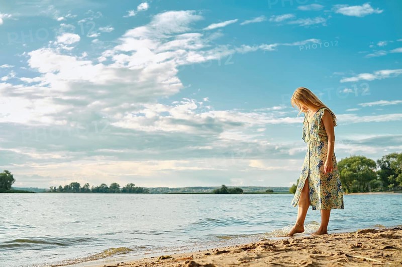 A woman wades along the sandy shore, wearing a colorful floral dress. The sun shines brightly, and fluffy clouds scatter across the vibrant blue sky, creating a tranquil atmosphere