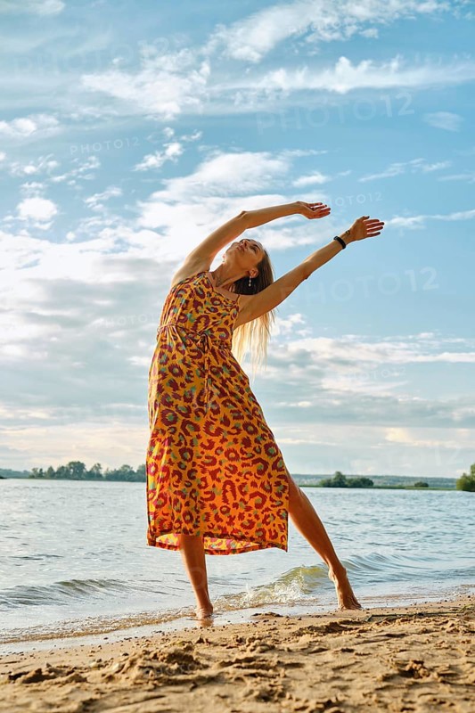 A joyful woman stretches gracefully on the sandy shore as the sun sets over the water. The vibrant dress she wears flows with her movements, capturing the serene beauty of the beach