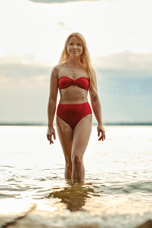 A woman with long hair walks confidently in shallow water, wearing a red bikini. The sun sets in the background, casting a warm glow over the beach. It is a serene summer evening by the lake