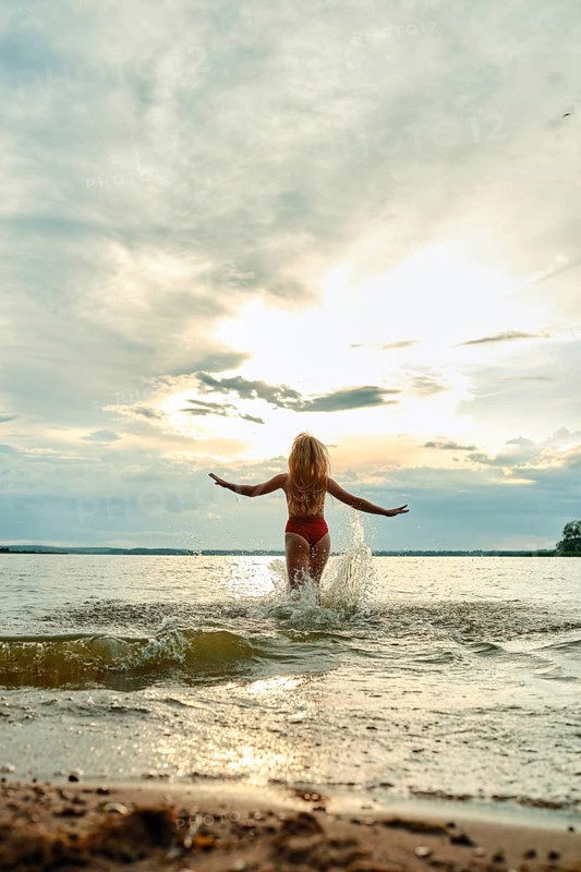 A woman in a red swimsuit wades into a shallow lake, arms outstretched, as the sun sets behind her. The sky is filled with soft clouds, and gentle waves lap at the sandy shore