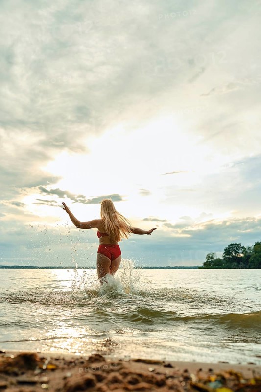 A woman in a red swimsuit plays in the shallow water of a lake. The sun sets behind her, casting a warm glow while she joyfully splashes and embraces the tranquil moment