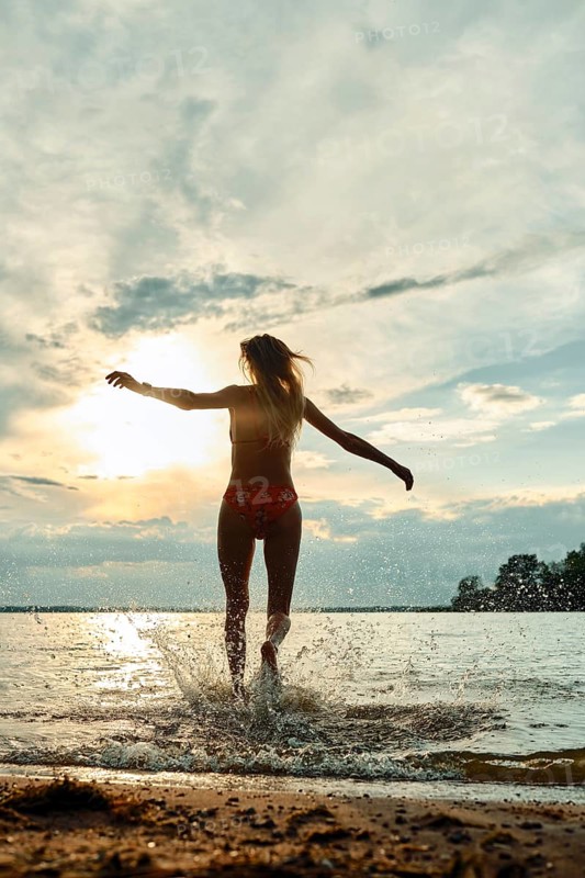 A woman wearing swimwear joyfully splashes through water at sunset. The sky displays vibrant colors, reflecting on the lake's surface. The tranquil atmosphere provides a sense of freedom and peace