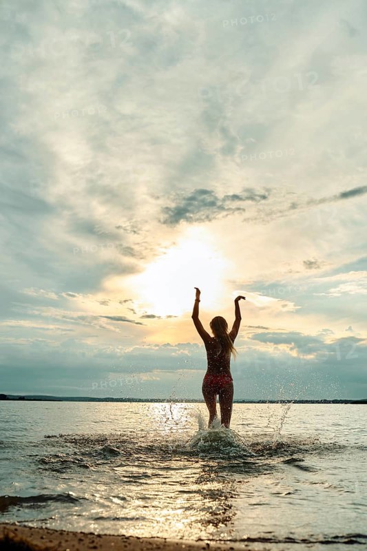 A person stands in the shallow water with arms raised, enjoying the sunset. The sky is filled with soft clouds, while the sun casts a warm glow over the tranquil water