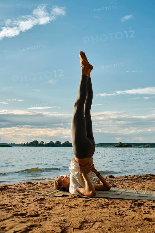 A woman performs shoulder blade stand with her legs raised high at a lakeside on a sandy shore. The sun sets behind her, casting a warm glow over the tranquil water and surrounding landscape
