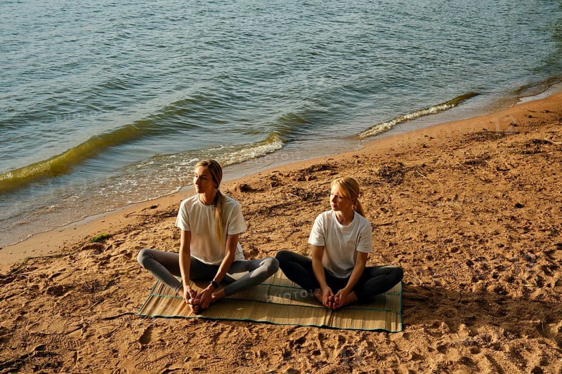 Two women are sitting on a mat by the water's edge and doing yoga poses as the sun begins to set. The sandy beach and gentle waves create a serene atmosphere for their practice
