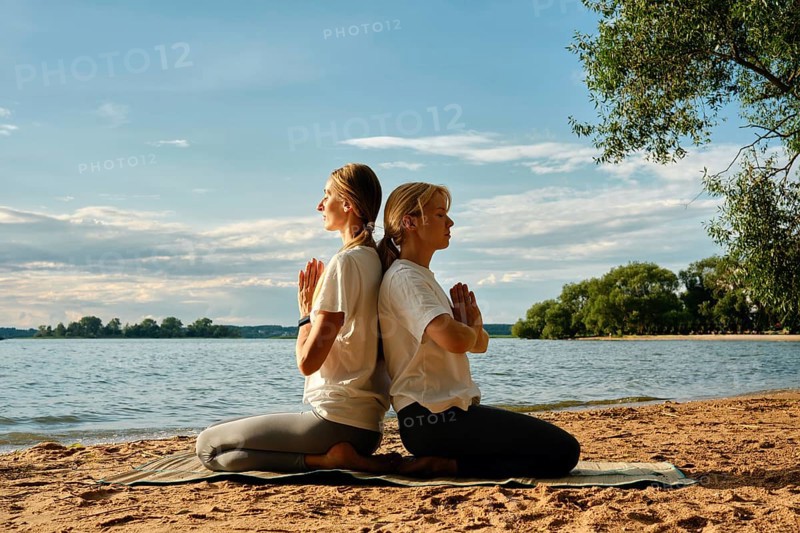 Two women sit back-to-back on a yoga mat, practicing mindfulness on a sandy beach by a tranquil lake. The warm evening light enhances their serene expressions as they find balance and connection