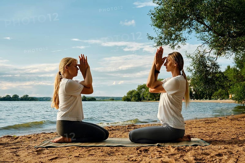 Two women sit on a mat in front of each other by the water, practicing yoga. They perform a meditative pose with hands raised, surrounded by nature as the sun sets behind them