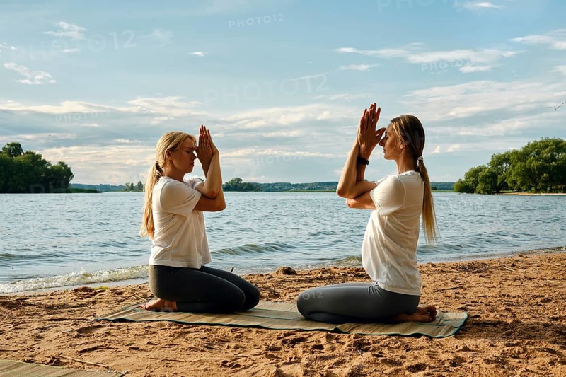 Two individuals are engaged in a yoga session on a sandy beach by a tranquil lake. The sun sets, casting soft colors across the sky. They perform a meditative pose sitting in front of each other