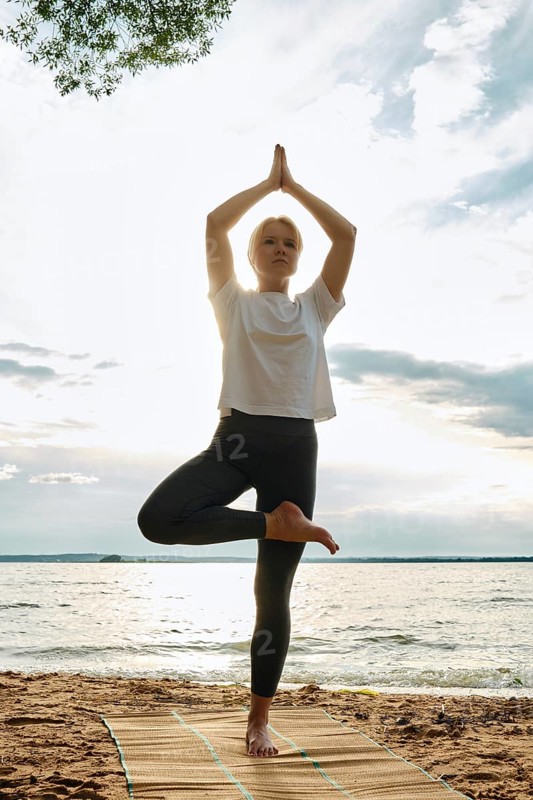 A woman performs yoga on a sandy beach, balancing in a tree pose with arms raised towards the sky. The sun sets behind, casting a warm glow over the peaceful water and clouds