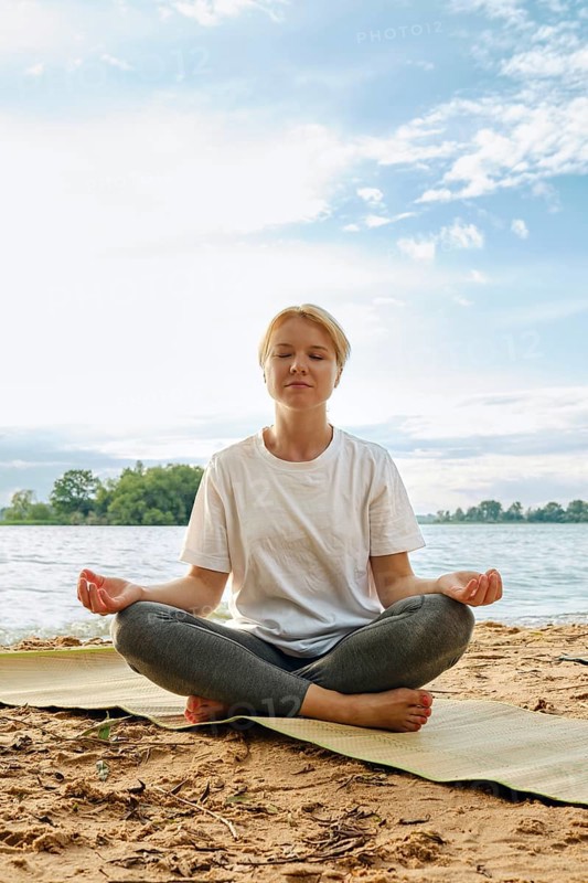 A woman sits cross-legged on a mat by the lake, eyes closed in meditation. The sun shines down on her as gentle waves lap at the shore, surrounded by lush greenery and blue skies