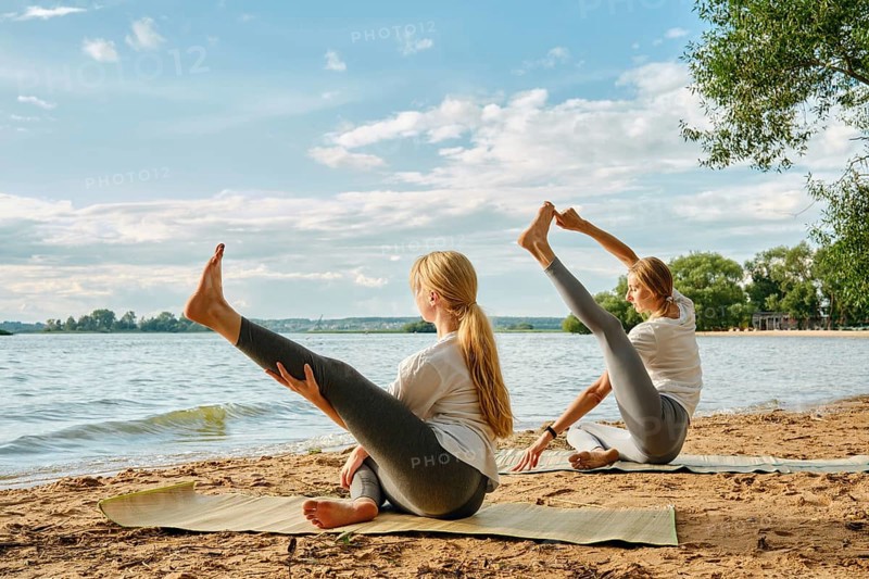 Two women practice yoga on mats by the lake, enjoying the sunny afternoon and calm waters. They showcase various poses while surrounded by trees and beautiful clouds in the sky