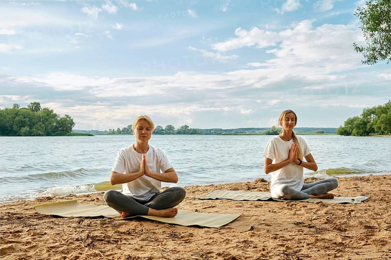 Two women are sitting cross-legged on yoga mats by the riverbank, practicing yoga with serene expressions. The sun shines brightly above, creating a peaceful atmosphere around them