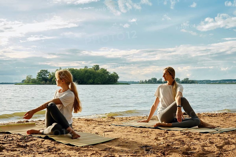 Two barefoot women practice yoga on mats on a sandy lakeshore at sunset