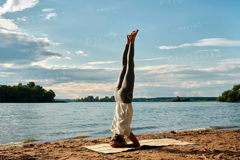 A woman performs a headstand yoga pose on a sandy beach beside a tranquil river. The sun sets in the background, casting warm hues across the sky and reflecting on the water