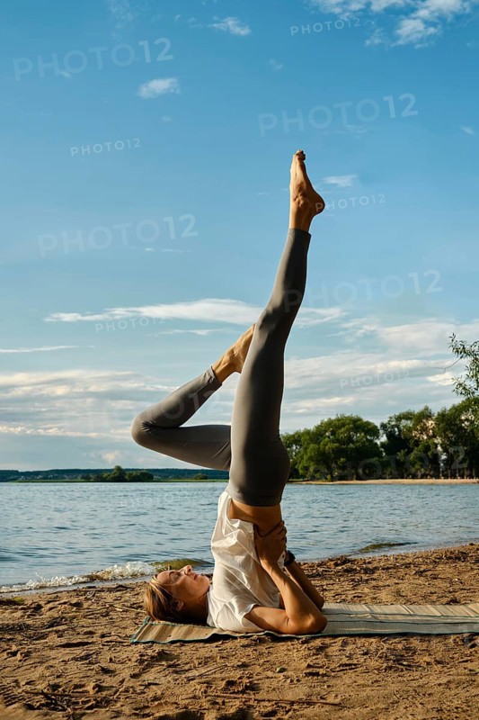 A dedicated individual performs a yoga pose on a sandy beach by the tranquil lake. The sun shines down, casting a warm glow, while lush trees line the water's edge, creating a peaceful atmosphere