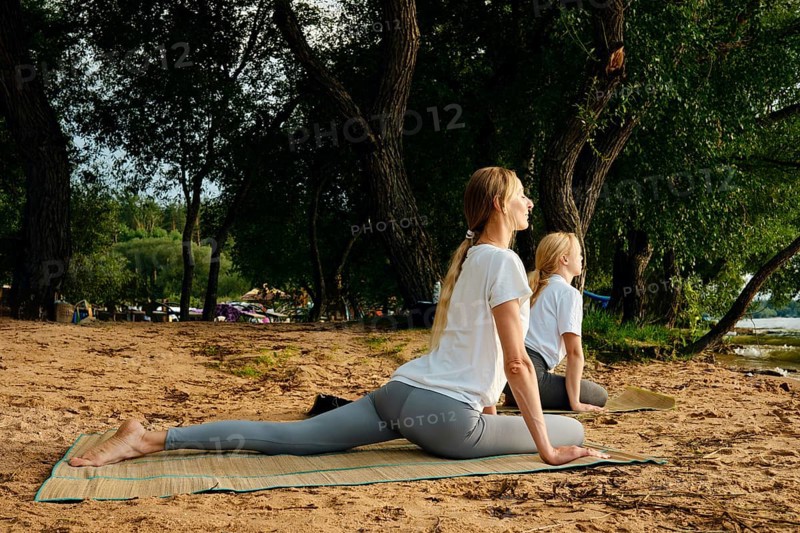 Two women are engaged in yoga poses on mats beside a tranquil lake at sunset. Surrounded by lush greenery, they focus on their breathing and balance, enjoying nature's calming atmosphere