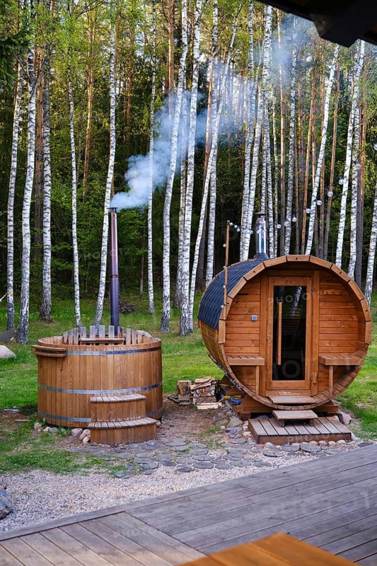 A cozy wooden sauna and hot tub surrounded by tall birch trees. Smoke gently rises from the chimney, creating a peaceful atmosphere in nature