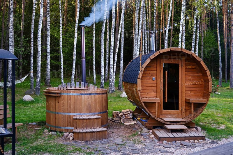 A wooden sauna and a hot tub are nestled in a lush forest. The hot tube emits smoke from its chimney, and a hammock nearby invites relaxation amid nature
