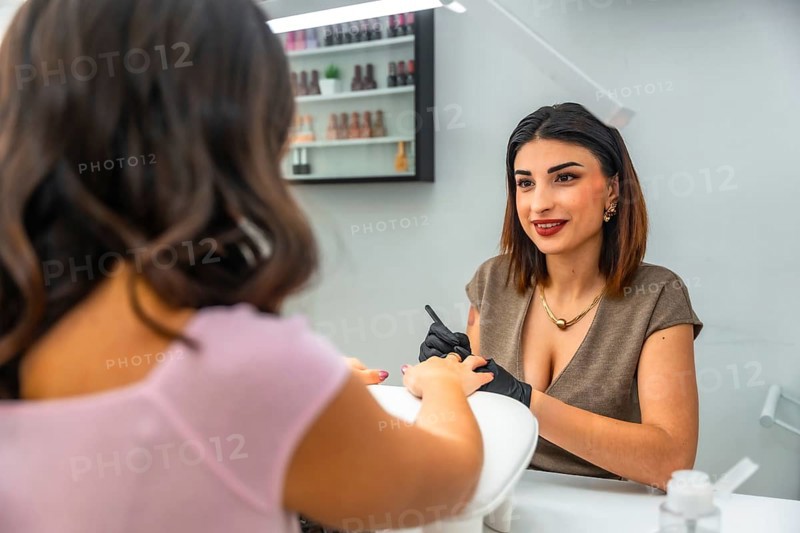 Smiling nail technician performing manicure on customer's fingernails, applying nail polish in a beauty salon