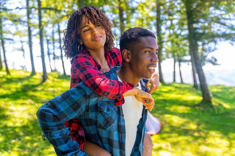 Young man giving piggyback ride to his girlfriend in forest during camping trip, enjoying summer vacation in nature