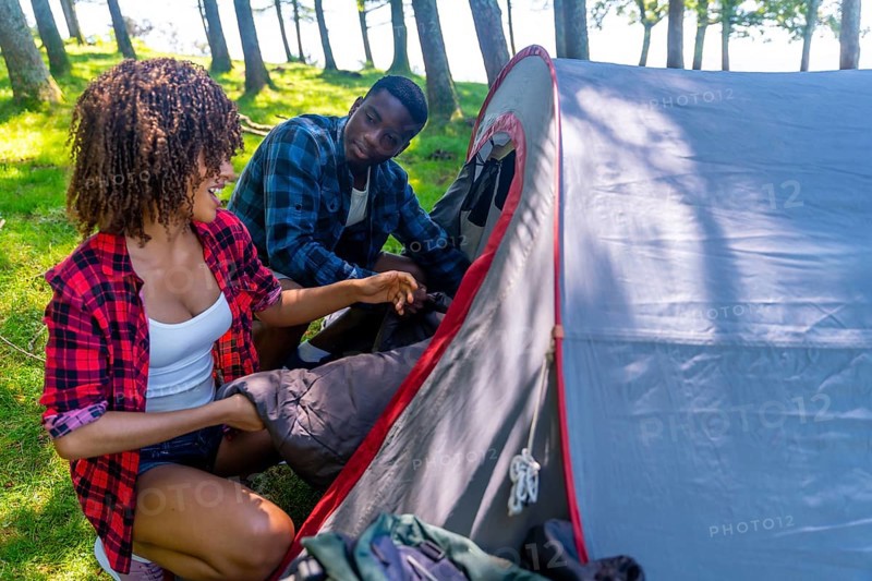 Young black couple enjoying the outdoors while setting up a camping tent in the forest, placing a sleeping bag inside with smiles