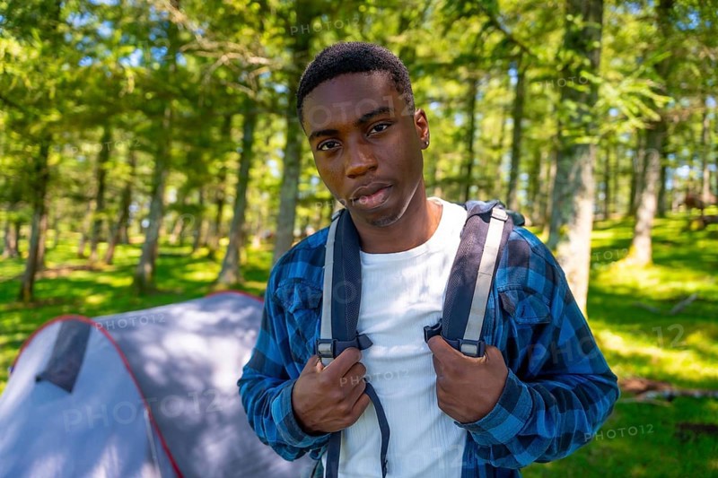 Young black man adjusts his backpack straps while standing in a forest clearing near his tent, enjoying a solo camping trip