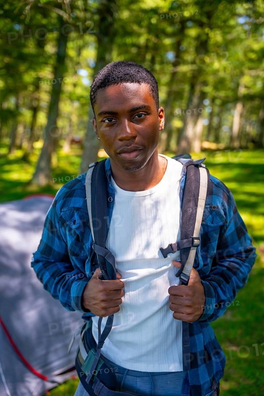 Young black man is wearing a backpack and standing in a forest near his tent, enjoying the peace of nature