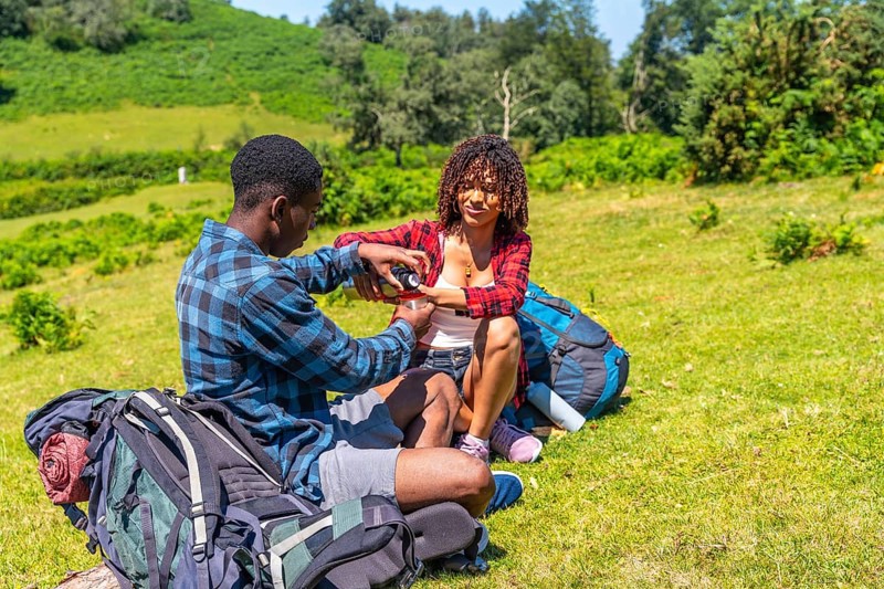 Young couple of hikers taking a break on a grassy hillside, sharing refreshments from a thermos during a sunny day hike