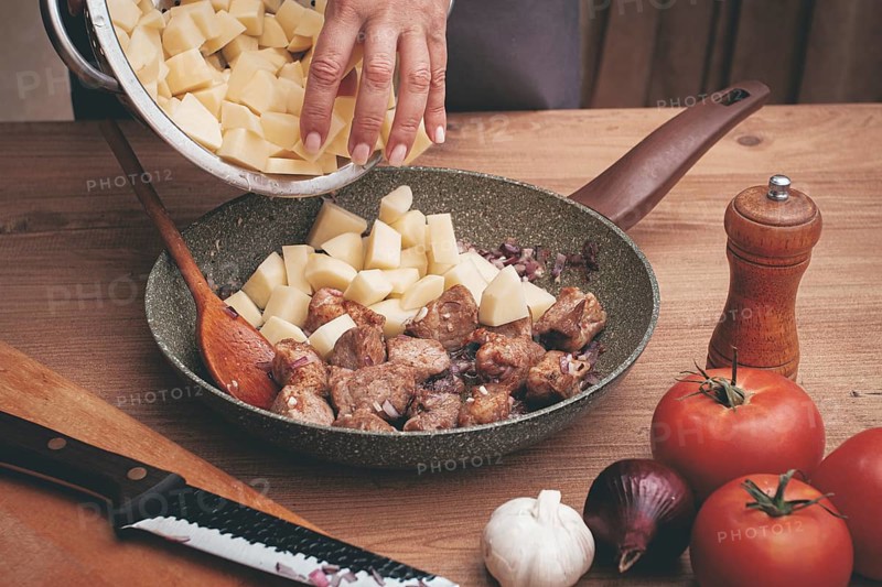 Potato slices are laid out in a frying pan, with browned meat, cooking stewed meat with potatoes