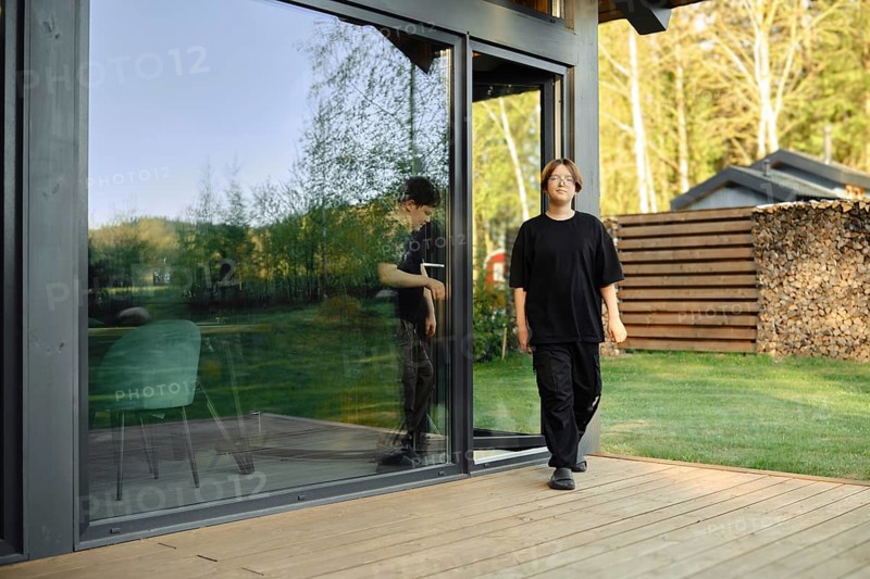 Two boys are walking out of a sleek, contemporary house showcasing large glass doors. The surrounding area features lush greenery and organized wood stacks, creating a peaceful atmosphere