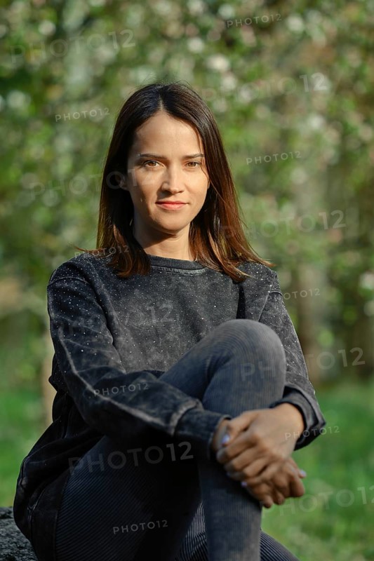 A young woman with long dark hair sits comfortably on a rock, wearing a casual sweater and leggings. She looks directly at the camera, with soft sunlight filtering through trees in the background